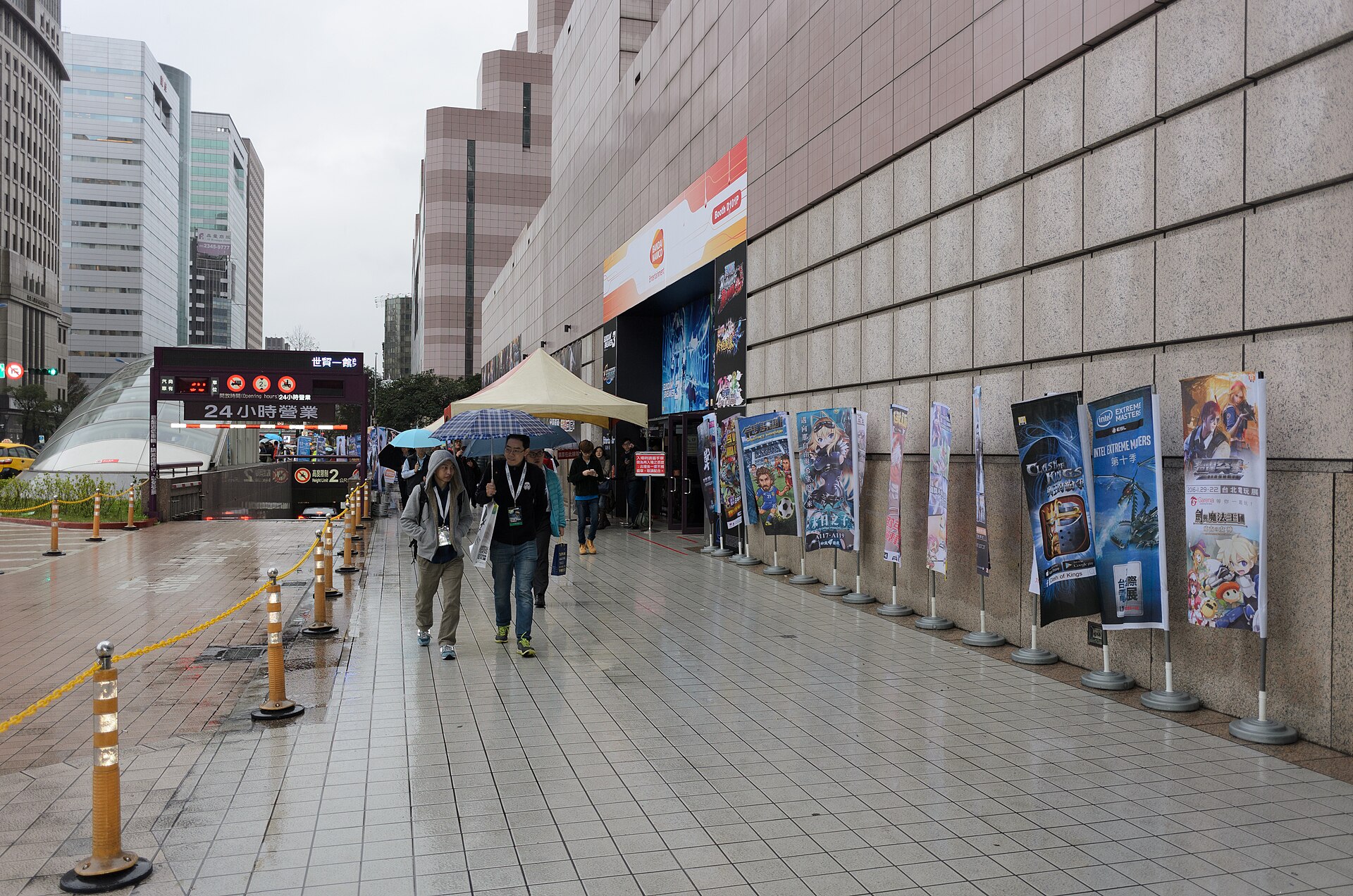 Visitors carry umbrellas along a wet sidewalk outside Taipei World Trade Center, beside exhibition banners and a metro entrance.