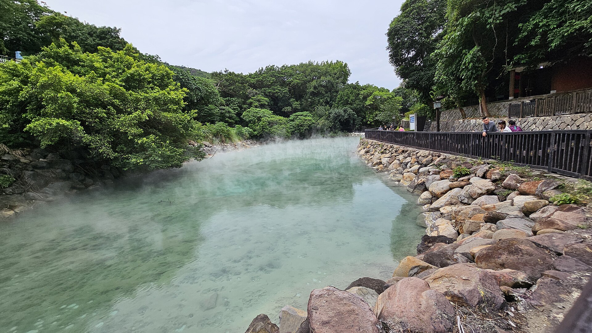 Visitors follow the railing beside steaming pale green water at Beitou Thermal Valley, with trees lining the opposite shore.