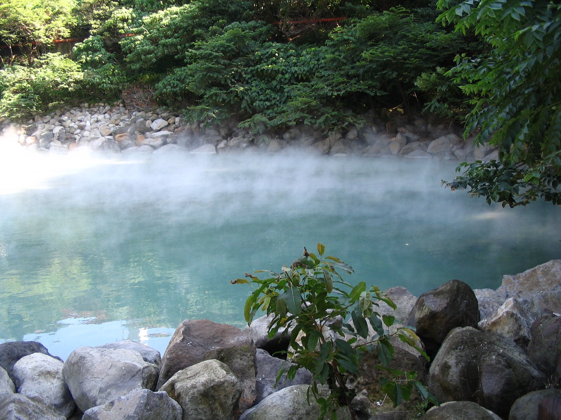 White steam hangs above turquoise water at Beitou Thermal Valley, framed by boulders and dense trees along the shore.