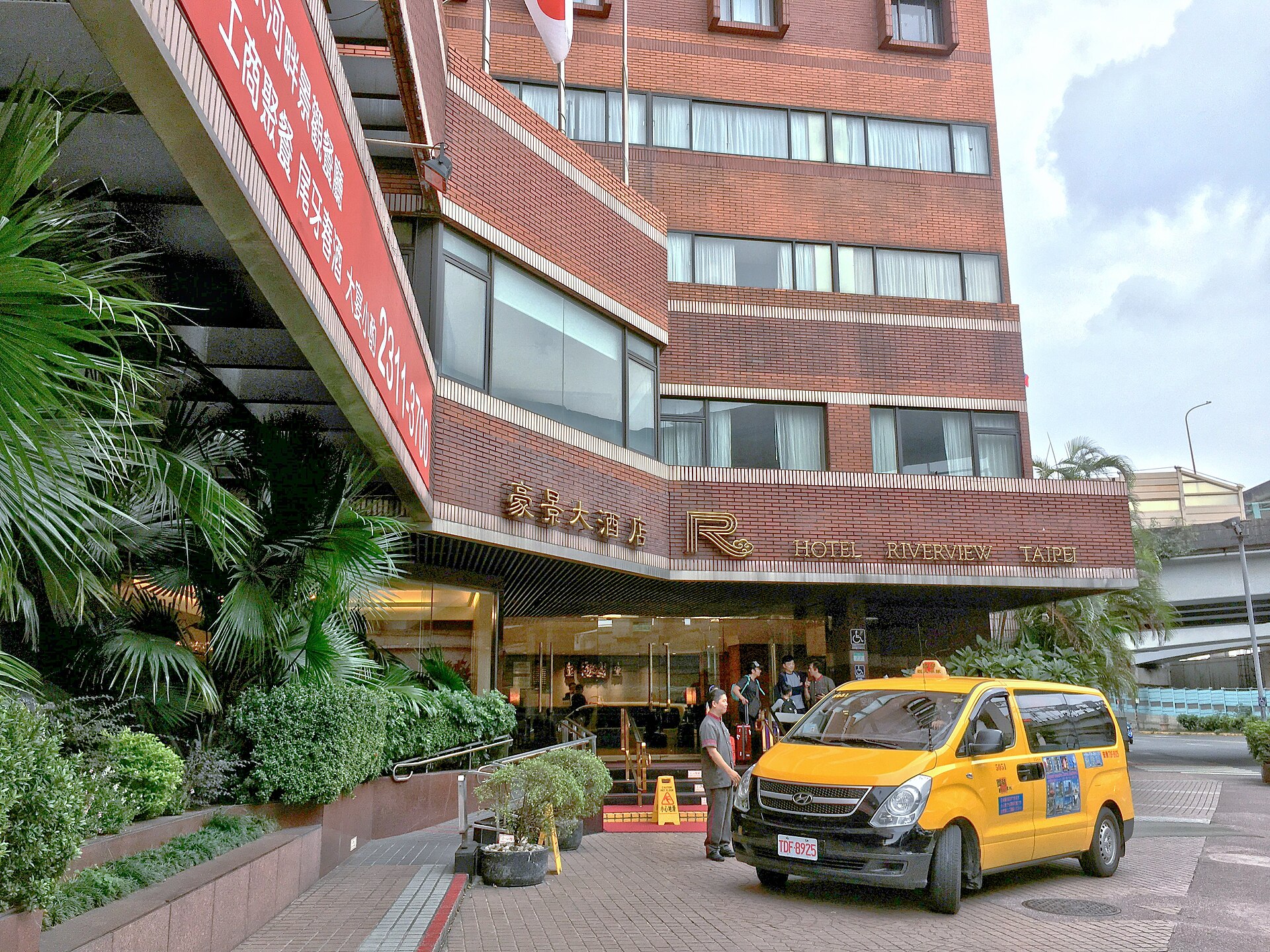 Yellow taxi van outside Hotel Riverview Taipei, with adults and luggage beneath the brick entrance canopy.