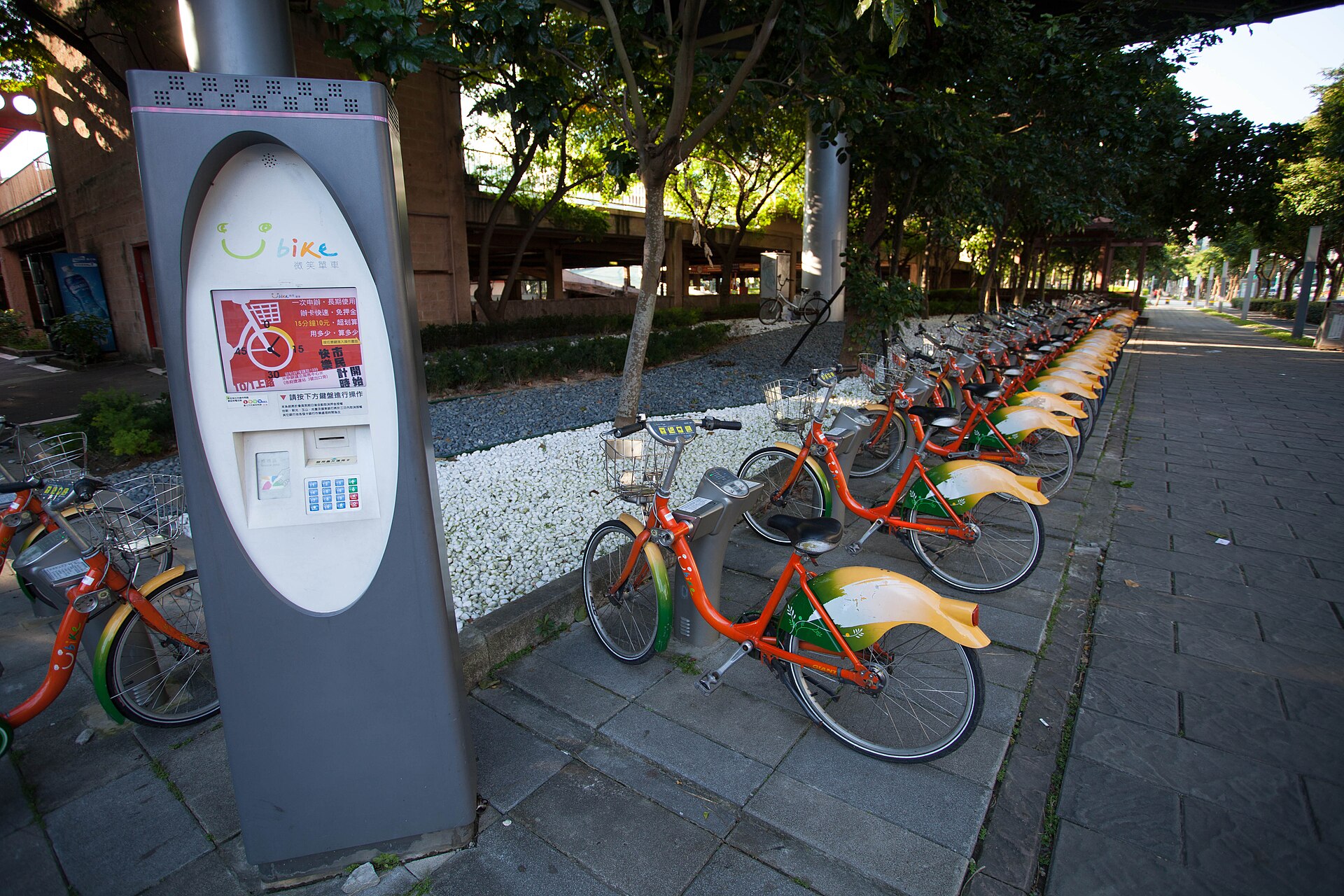 A YouBike rental kiosk and rows of orange shared bicycles line a shaded sidewalk in Xinyi, Taipei.
